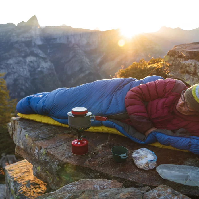 Campeur en bivouac profitant d'un lever de soleil en montagne, confortablement installé dans un sac de couchage, avec un réchaud et des provisions à ses côtés.