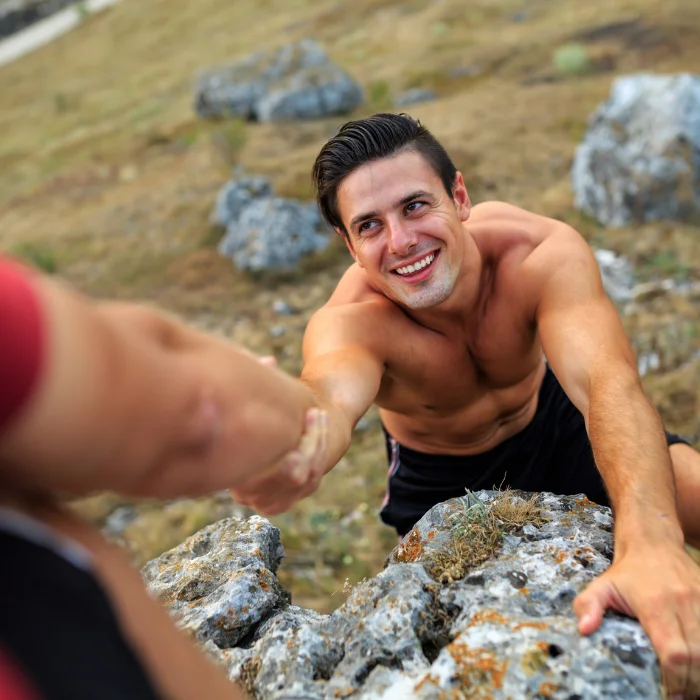 Homme souriant recevant de l'aide pour grimper lors d'une aventure en plein air, illustrant l'esprit de coopération et de solidarité en bivouac. Un moment de camaraderie et de dépassement de soi.