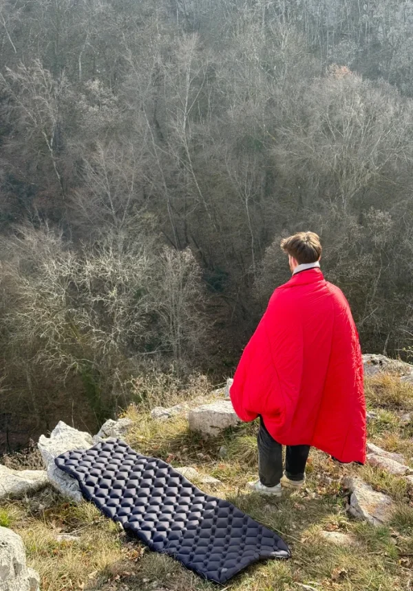 personne observant debout au bord de la montagne avec un duvet sur ses épaules et à coté un matelas gonflable de trek