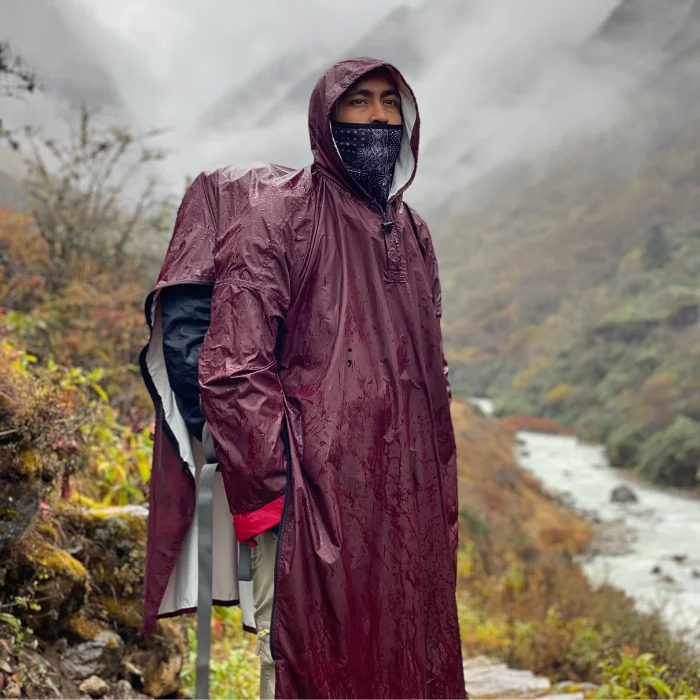 Homme équipé d’un poncho bordeaux avec capuche, en randonnée sur un sentier de montagne sous la pluie.
